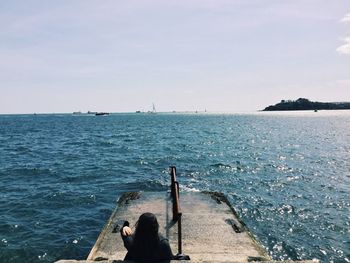 Rear view of woman sitting on pier overlooking sea against clear sky