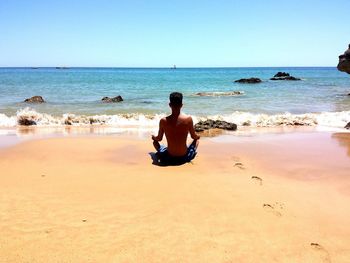 Rear view of shirtless young man practicing yoga on shore at beach against clear sky