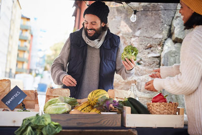 Smiling man buying fresh broccoli from female market vendor at vegetable stall