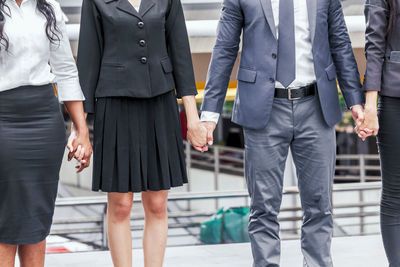 Midsection of colleagues holding hands while standing on elevated walkway against buildings in city