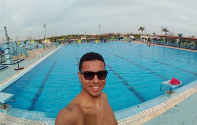 Portrait of shirtless young man wearing sunglasses while standing at poolside against sky