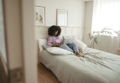 Young woman using laptop sitting on bed in bedroom