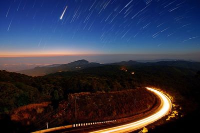 Illuminated country road amidst mountain against shooting stars during night