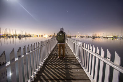 Rear view of man standing on footbridge over sea at night