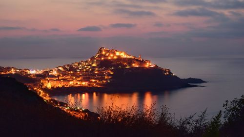 Illuminated city by sea against sky during sunset