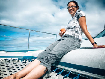 Low angle portrait of smiling woman sitting on railing against sky