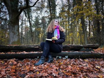 Young woman sitting down on a wooden bench in the forest during autumn and reading