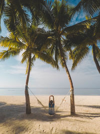 Rear view of woman sitting on hammock at beach against sky