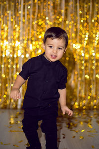 Boy in black clothes at christmas standing in a room on a background of gold tinsel on the wall