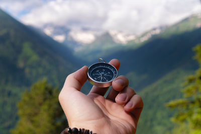 A hand with a compass against the backdrop of epic snow-capped mountains with clouds and a forest at