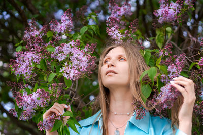 A woman is standing in front tree with purple flowers. she is wearing blue shirt and a necklace