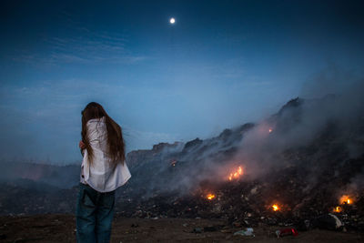 Rear view of woman standing by burning garbage at junkyard against sky