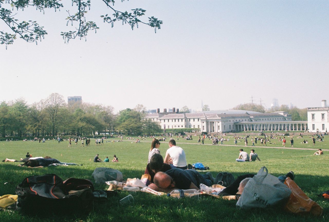 PEOPLE SITTING ON GRASSLAND AGAINST BUILDINGS