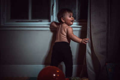 Rear view of shirtless baby boy standing by window at home