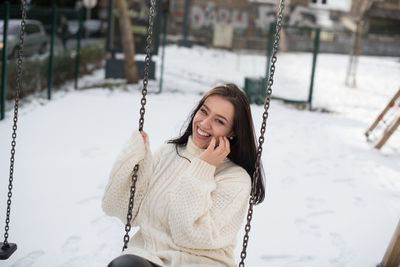 Portrait of happy young woman swinging in snow covered playground
