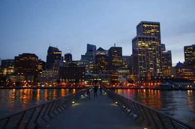 Illuminated buildings by river against sky in city at dusk