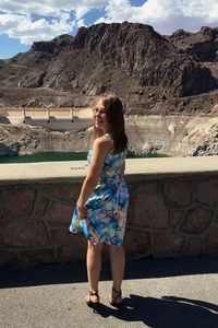 Smiling woman wearing blue dress standing by retaining wall against rocky mountains