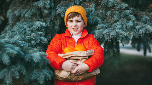 A cute boy in a bright orange jacket and a yellow hat holds a large package with oranges 