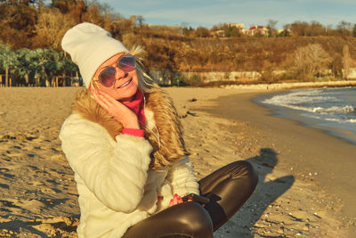 A woman in a fur coat and hat sits on a sandy beach and listens to music on a smartphone. 