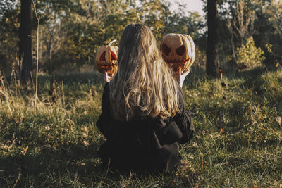 Rear view of woman holding jack o' lanterns while sitting on grassy field in forest during halloween