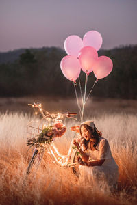 Woman with balloons sitting on illuminated bicycle on field against sky at sunset