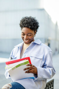 Positive african american female student with short hair reading information in documents while sitting on street against blurred background in city