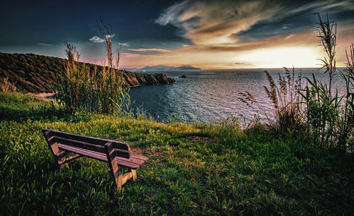 Empty bench on grass by sea against sky during sunset