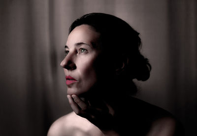 Shirtless mature woman with hand on chin looking away against curtain at home