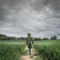 Rear view of a woman wearing a parka coat walking along a track in a wheat field with a stormy sky.