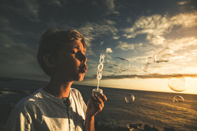 Close-up of cute boy blowing bubbles against sky during sunset