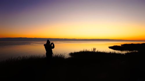 Silhouette woman photographing lake against sky during sunset