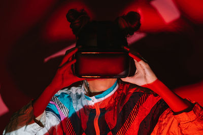 Young female in vr headset exploring virtual reality while standing on red background in studio with dim light