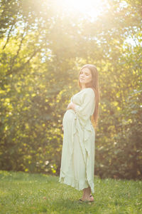 Young pregnant woman holds big belly, green trees, sunlight on background. brown haired female