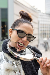 Portrait of woman in sunglasses biting mirror against building