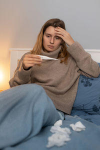 Young woman sitting in bed, touching head and looking on thermometer. girl measures body temperature