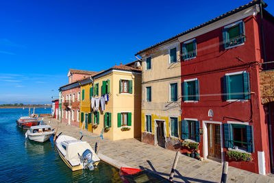 Boats moored on canal by houses against clear blue sky