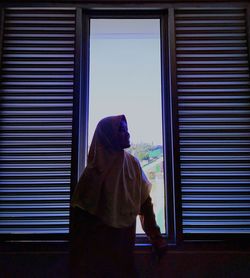 Young woman wearing hijab while standing by window at home