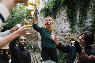 Cheerful woman with mouth open toasting drinks with friends during dinner party in back yard