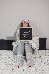 Smiling woman with merry christmas text sitting on bed at home