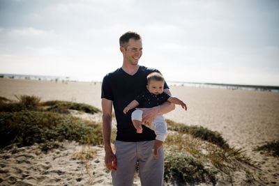 Father casually standing on beach holding infant son