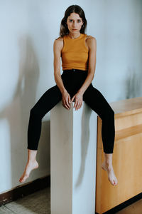 Full length portrait of young woman sitting by wall