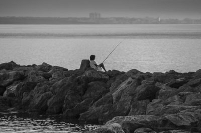 Rear view of man fishing while sitting on rocks against sea