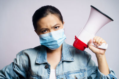Portrait of young woman wearing flu mask holding megaphone standing against gray background
