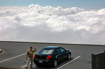 Man standing by car at parking lot against cloudy sky