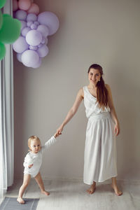 Girl child stands next to her mother at the window in white clothes at home in winter