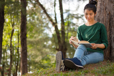 Full length of young woman reading book while leaning on tree at park