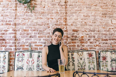 Smiling young woman talking over smart phone while sitting against brick wall in cafe