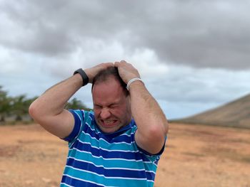 Angry man with hand in hair standing on field against cloudy sky