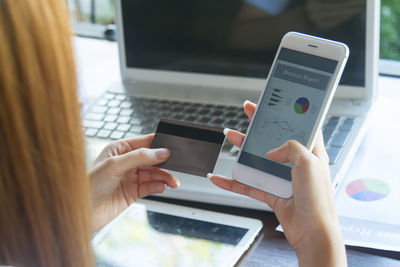 Cropped image of businesswoman holding credit card while analyzing graph in mobile phone with laptop and digital tablet on table