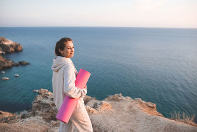 Smiling woman hold yoga mat wear sportswear hoodie and pants stand on top of rock over sea